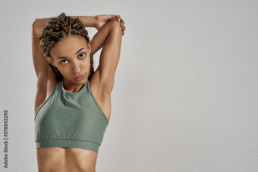 Fototapeta premium Good warm up routine. Young mixed race woman in sportswear stretching her arms before fitness workout and looking at camera while standing isolated over grey background