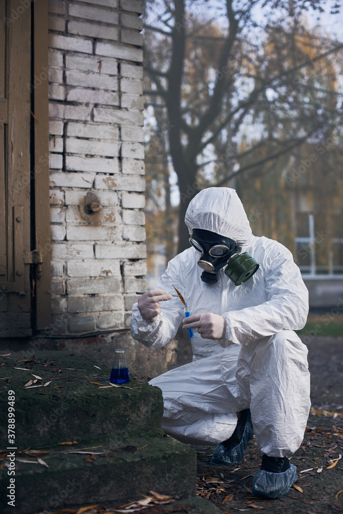 Researcher in protective coverall and a gas mask, making tests using laboratory tools and local samples near abandoned buildings