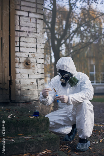 Researcher in protective coverall and a gas mask, making tests using laboratory tools and local samples near abandoned buildings