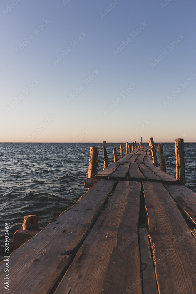Fototapeta premium Wooden pier at the sunset. Evening sky over sea with footbridge. Calm evening landscape. Empty pier at seaside with copy scape. Pier in warm sunset light. Horizon over sea. Marine travel. 