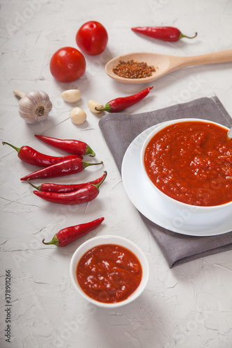 Spicy harissa sauce in a white porcelain dish with a sof eroy linen napkin, chili peppers, garlic and tomatoes on a white concrete background. vertically
