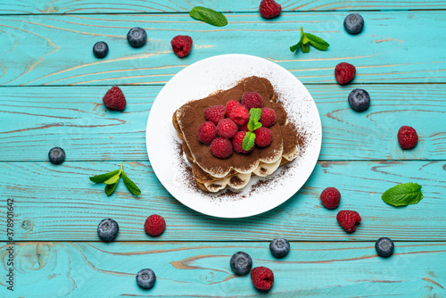portion of Classic tiramisu dessert with raspberries and blueberries on blue wooden background