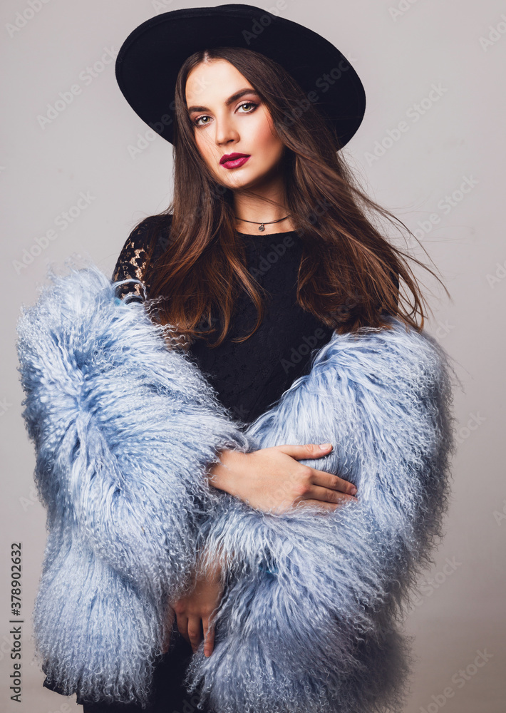 Close up indoor studio fashion portrait of pretty young model in stylish winter fluffy coat and black hat posing . Evening bright make up.