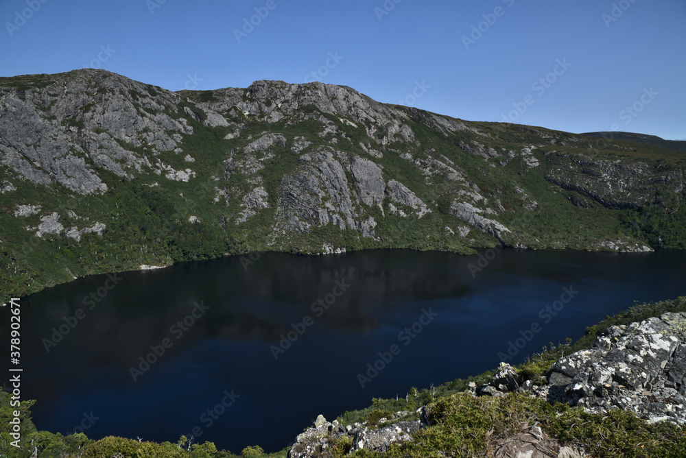 High angle view on Crater Lake. Deep blue water surrounded by rocky mountain cliffs and green forest ,The Overland Track, Tasmania, Australia