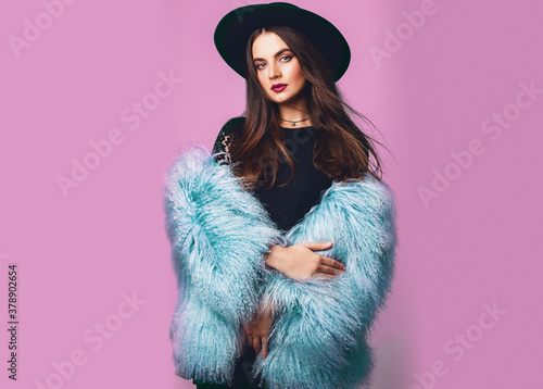 Close up indoor studio fashion portrait of gorgeous  woman in stylish winter fluffy  blue coat and black hat posing on bright pink background .