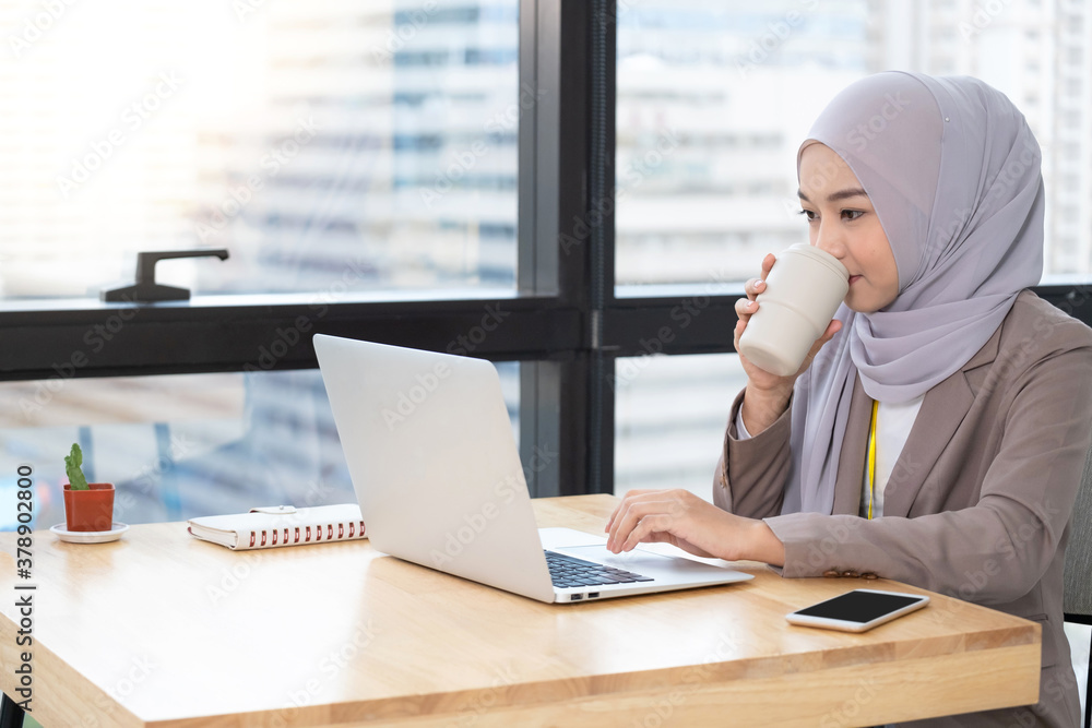 Confident Asia Muslim (Islam) businesswomen executive working in the modern office drinking a cup of coffee and looking on a laptop and sit beside windows. Work concept of diversity of culture.. Confident Asia Muslim (Islam) businesswomen executive working in the modern office drinking a cup of coffee and looking on a laptop and sit beside windows. Work concept of diversity of culture..