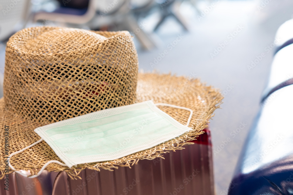 Defocus focus and blurred shot of surgical face mask, together with straw hat and travel luggage inside waiting seat area of Airport terminal. New normal lifestyle for traveler after Coronavirus. - obrazy, fototapety, plakaty 