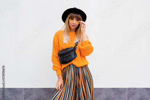 Slika na platnu Happy smiling brunette  woman  standing on white background in stylish orange sweater and multicolor stripe culotte , oversize orange knitted sweater, trendy bum bag and hat