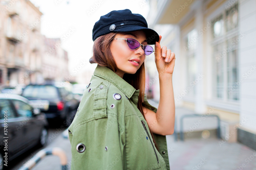 Playful woman posing outdoor . Wearing trendy purple retro glasses , black cap . Close up portrait . Windy hairs. Lifestyle image.