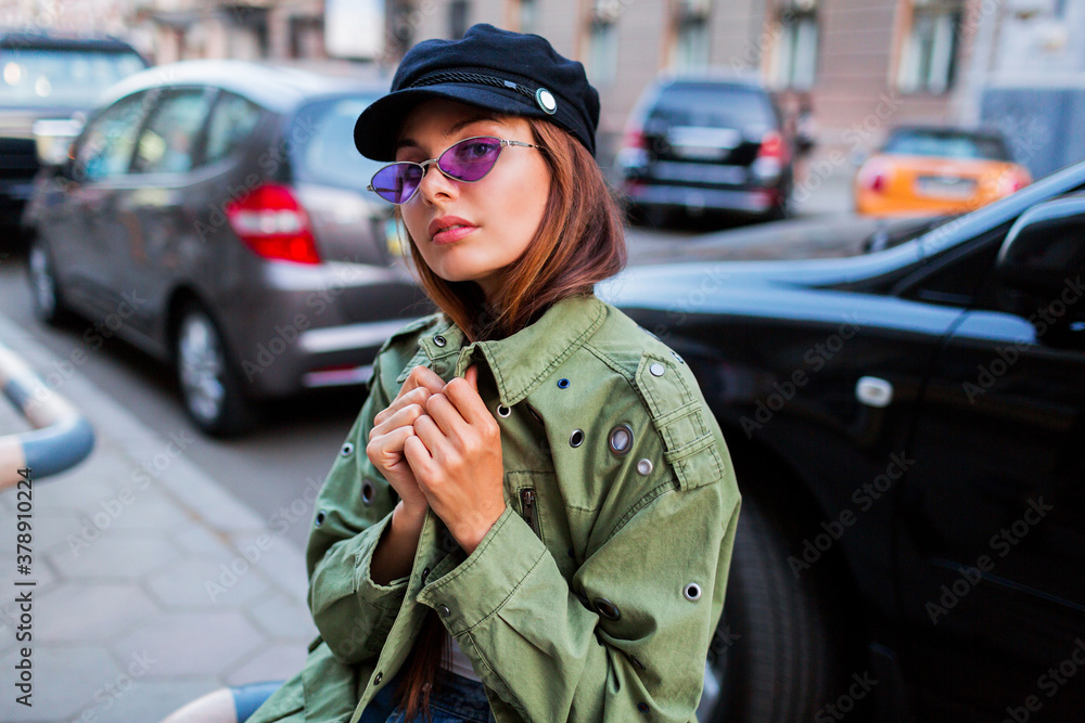 Fototapeta premium Beautiful girl looking at camera. Close up portrait of fashionable lady in green jacket sitting near road outdoor. Car parking background.
