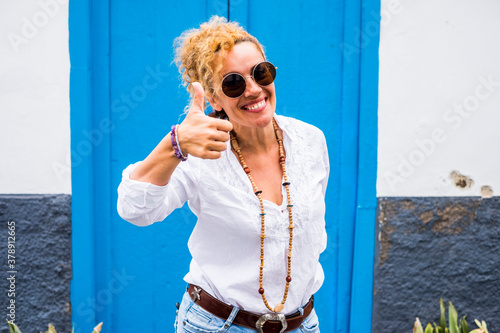 Cheerful and beautiful ok sign caucasian woman portrait with blue door home in background - concept of new house real estate business with happy people smiling and posing outdoor