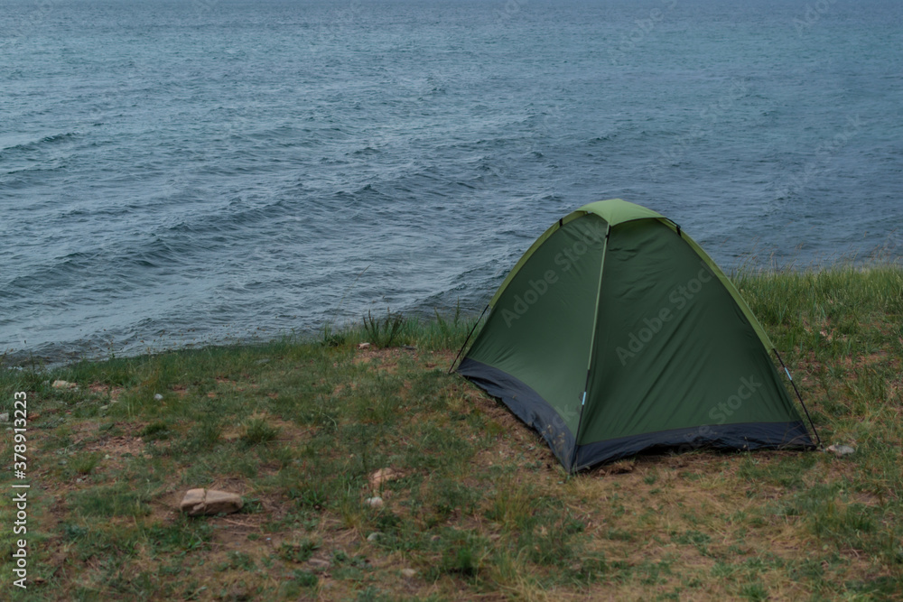 lonely green small tent stands on grass shore of blue Lake Baikal, sea waves and ripples, summer, overcast