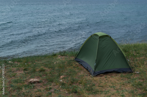 lonely green small tent stands on grass shore of blue Lake Baikal, sea waves and ripples, summer, overcast