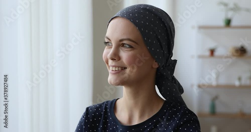 Head shot 25s young woman wear headkerchief on bald head after chemotherapy therapy smile look out the window standing indoor, welcoming day begin new life after recovery of oncology disease concept photo