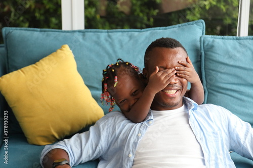 African American father playing peek a boo with his little daughter while spending happy time together at home on sofa couch photo
