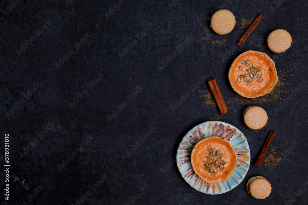 autumn orange creamy pumpkin pies for halloween with seeds, ceramic plate, cinnamon sticks, several macaroon cakes on a black background, top view, place for text, flat lay autumn orange creamy pumpkin pies for halloween with seeds, ceramic plate, cinnamon sticks, several macaroon cakes on a black background, top view, place for text, flat lay