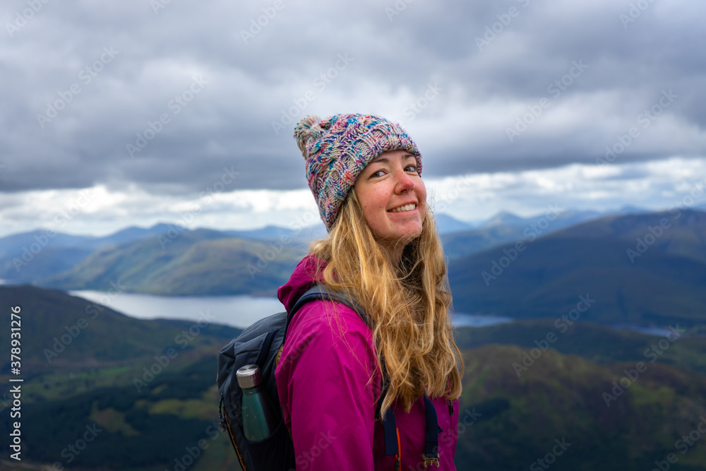 Woman smiling and happy while hiking Ben Nevis mountain, Scotland, UK with a winter hat and a beautiful backdrop of mountains and lakes