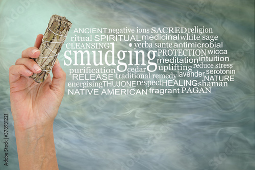 Words associated with smudging - female hand holding up a white sage smudge stick beside a SMUDGING word tag cloud against a green grey ethereal smoky background 
 photo