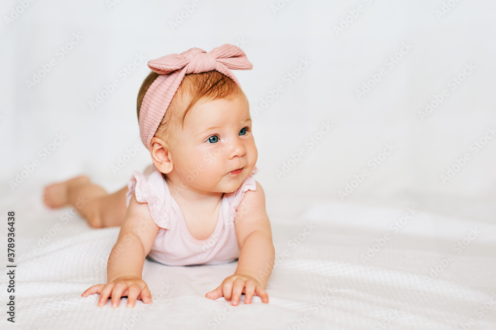 Portrait of adorable red-haired baby lying on belly, white background, wearing pink body and headband, looks to the side Portrait of adorable red-haired baby lying on belly, white background, wearing pink body and headband, looks to the side