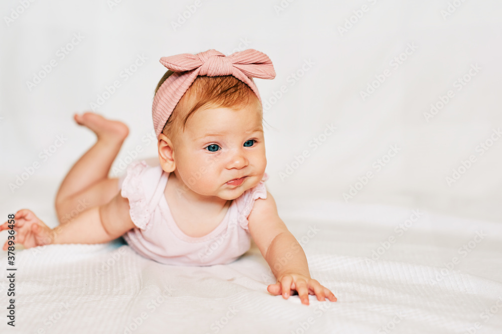 Portrait of adorable red-haired baby lying on belly, white background, wearing pink body and headband, infant with funny facial expression Portrait of adorable red-haired baby lying on belly, white background, wearing pink body and headband, infant with funny facial expression