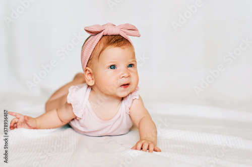 Portrait of adorable red-haired baby lying on belly, white background, wearing pink body and headband, looks to the side