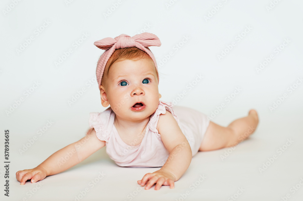 Portrait of adorable red-haired baby lying on belly, white background, wearing pink body and headband Portrait of adorable red-haired baby lying on belly, white background, wearing pink body and headband
