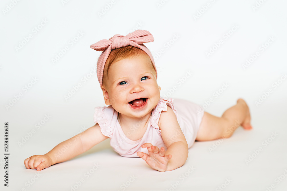 Portrait of adorable red-haired baby lying on belly, white background, wearing pink body and headband Portrait of adorable red-haired baby lying on belly, white background, wearing pink body and headband