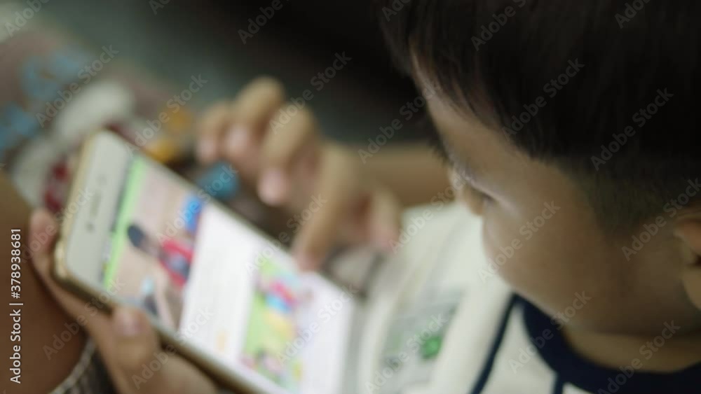 A three-year-old Asian boy is sitting on armchair and staring at social media on his parent's mobile phone joyfully. A three-year-old Asian boy is sitting on armchair and staring at social media on his parent's mobile phone joyfully.