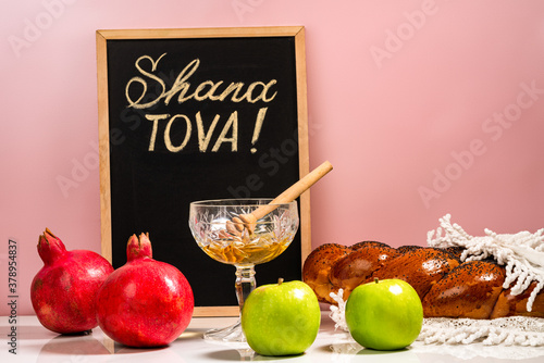 Hebrew inscription Good year (Shanah tovah) on a chalk board. Apples, pomegranates, honey and wicker challah on a wooden table.