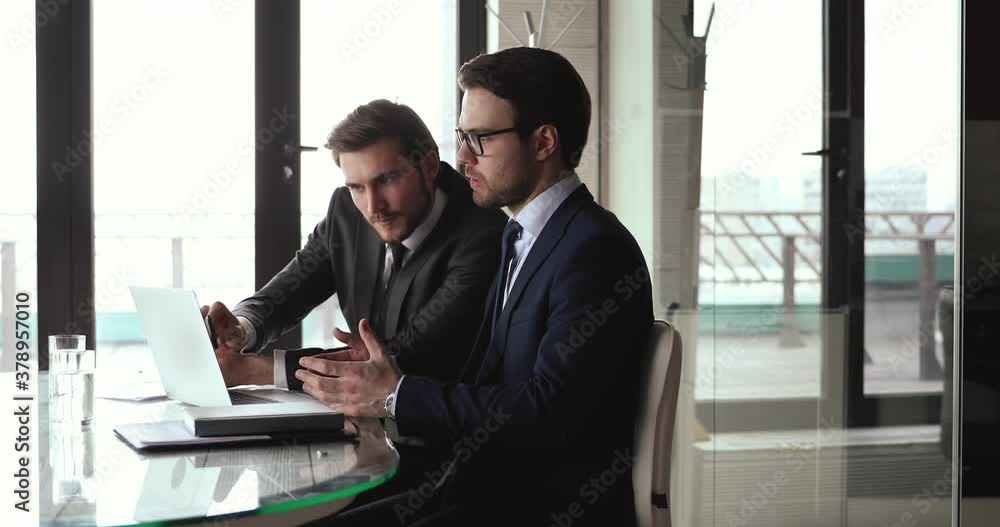 Two focused businessmen in formal wear discussing project presentation on computer, shaking hands after making agreement. Financial advisor handshaking with client after presenting profitable offer. Two focused businessmen in formal wear discussing project presentation on computer, shaking hands after making agreement. Financial advisor handshaking with client after presenting profitable offer.