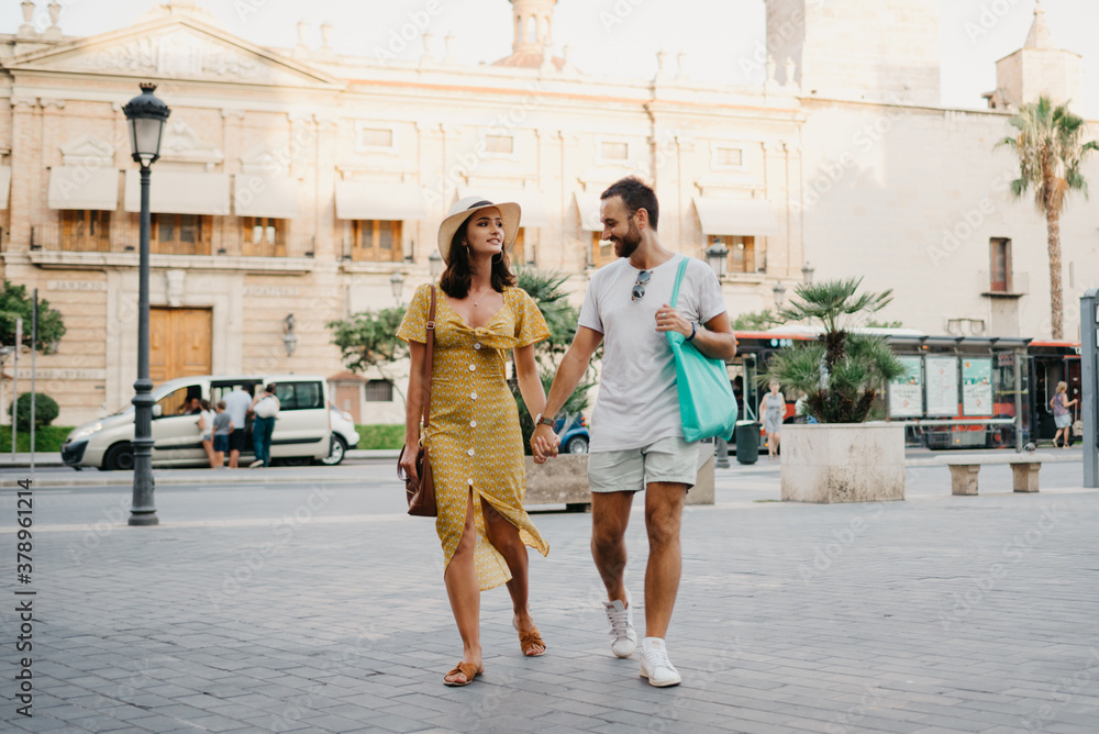 Girl in a hat and a dress with a plunging neckline and her boyfriend with a beard and sunglasses are relaxing holding each other's hand in the old town. A couple of tourists on the sunset in Valencia.