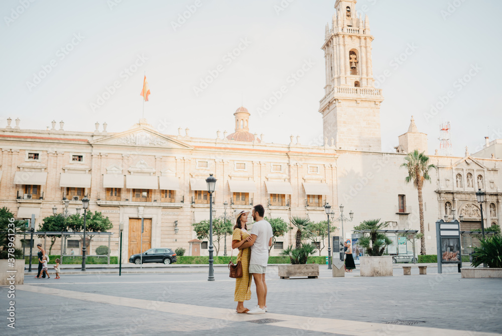 Obraz premium A girl in a hat and a dress with a plunging neckline is dancing with her boyfriend with a beard and sunglasses in the old town. A couple of tourists on the sunset in Valencia.