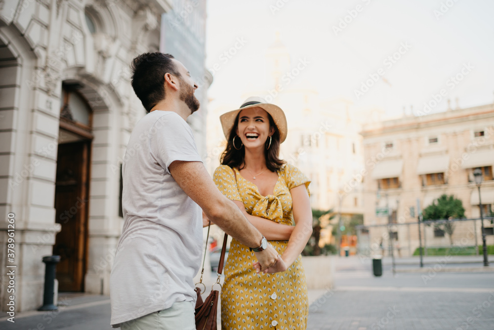 A laughing girl in a hat and a yellow dress with a plunging neckline is dancing with her boyfriend with a beard and sunglasses in the old town. A couple of tourists on the sunset in Valencia.