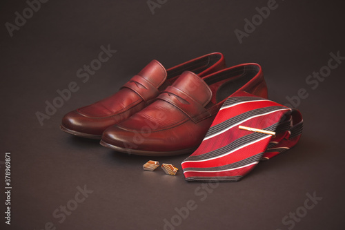Stylish red striped rolled necktie, gold colored tie pin, cufflinks and fashionable brown men's shoes on dark background. Selective focus.