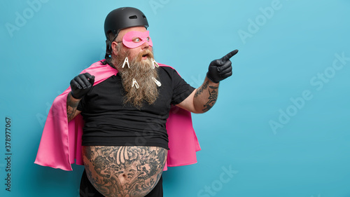 Photo Shocked superhero points at blank space wears pink mantel and mask shows something astonishing isolated over blue background wears clothespins on thick beard