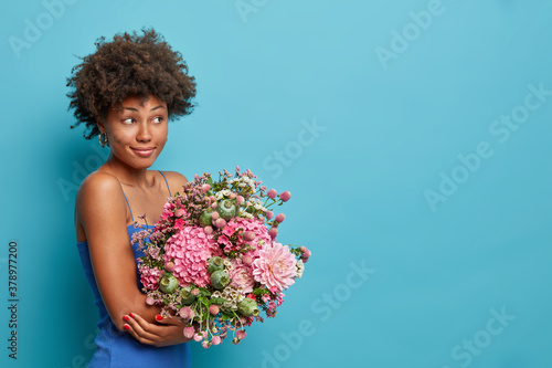 Smiling woman with Afro hair holds beautiful bouquet of flowers has date with boyfriend looks mysterious aside poses against blue studio background copy space for your promotion. Spring time