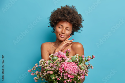 Tender gorgeous female model touches jawline gently stands with closed eyes bare shoulders enjoys soft skin holds beautiful bouqet of flowers isolated on blue background. Natural beauty concept