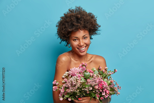 Indoor shot of glad smiling woman has healthy skin and natural curly hair poses bare shoulders with present bouquet of flowers against blue background enjoys pleasant aroma. Holiday concept.