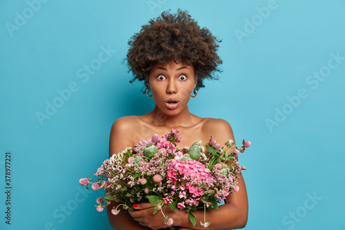 Horizontal shot of scared dark skinned woman with curly hair stares at camera keeps big nice festive bouquet of flowers over naked body isolated on blue background. Womens day spring time concept