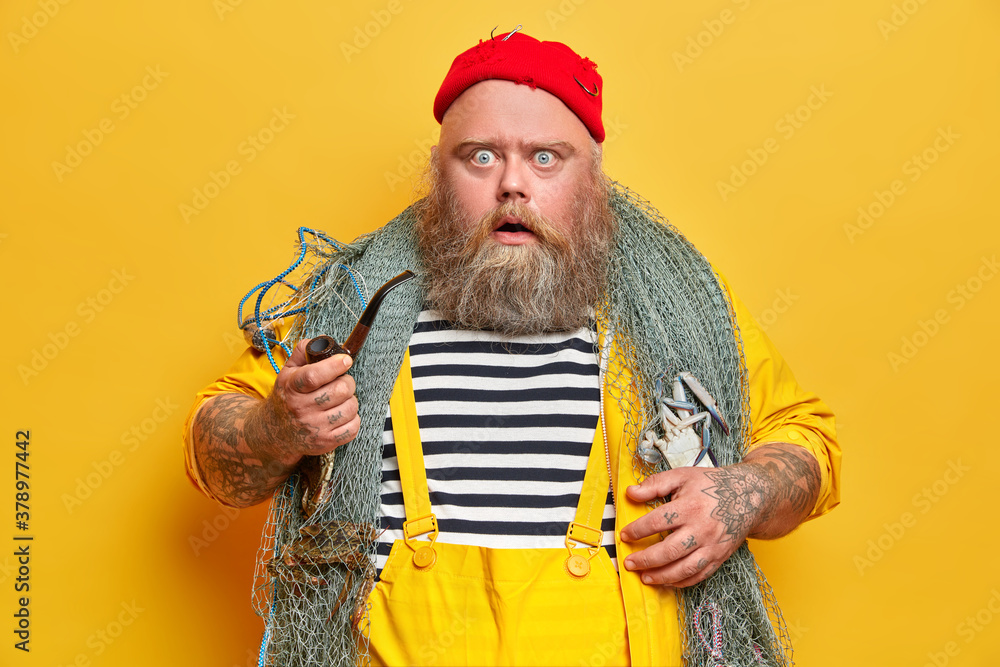 Plump bearded professional man mariner stares at camera with displeased shocked expression holds smoking pipe and goes fishing isolated on yellow background. Fatso seaman stunned to see storm at ocean - obrazy, fototapety, plakaty 