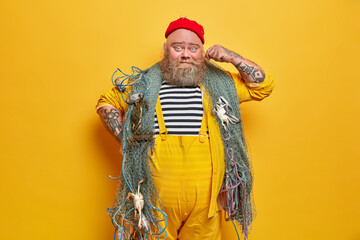Photo of bearded sailor man curls mustache and poses indoor with fishing gears poses against yellow background. Professional boatswain poses on deck. Mariner in overalls enjoys sea adventure