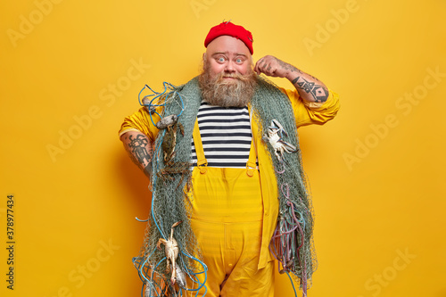 Photo of bearded sailor man curls mustache and poses indoor with fishing gears poses against yellow background. Professional boatswain poses on deck. Mariner in overalls enjoys sea adventure