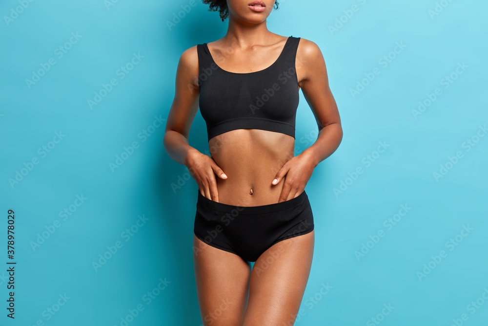 Sporty dark skinned woman demonstrates results after fitness training dressed in black cropped top and panties cares about body isolated on blue background - obrazy, fototapety, plakaty 