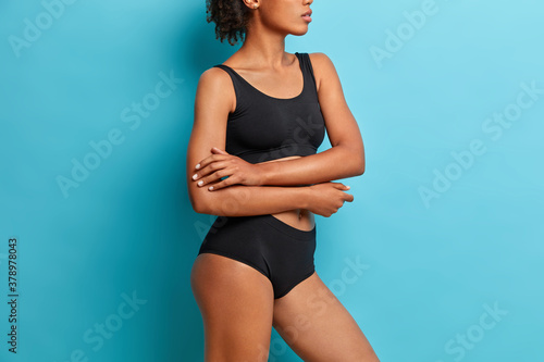 Sideways shot of sexual young woman stands with arms crossed over body dressed in black underwear has healthy skin poses against blue background