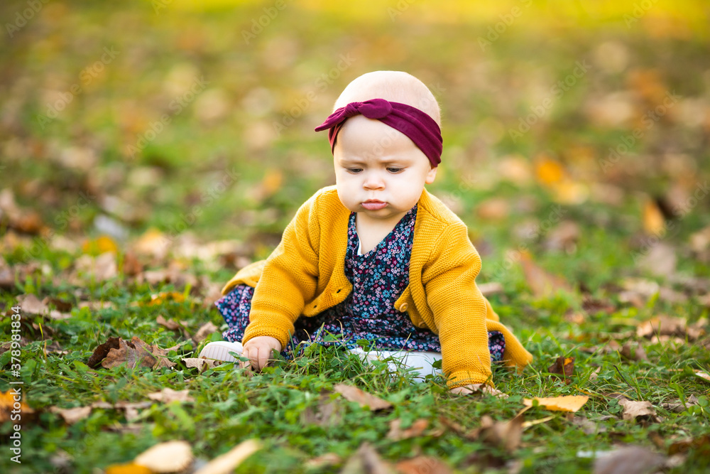 Baby girl in yelloy jacket and red headband seating on the grass, playing in the autumn leaves