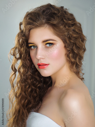 Glamour portrait of beautiful woman model with professional fresh trendy makeup shiny highlighter on skin, sexy glossy lips romantic curly hairstyle natural light light background
