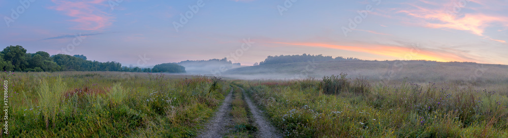 Rural panoramic landscape with country road passing through meadows at foggy morning