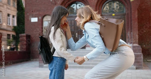 Beautiful happy Caucasian woman talking with little cute girl before classes at schoolyard. Joyful pretty junior student hugging her mom and running to school. Lovely family. Study concept photo