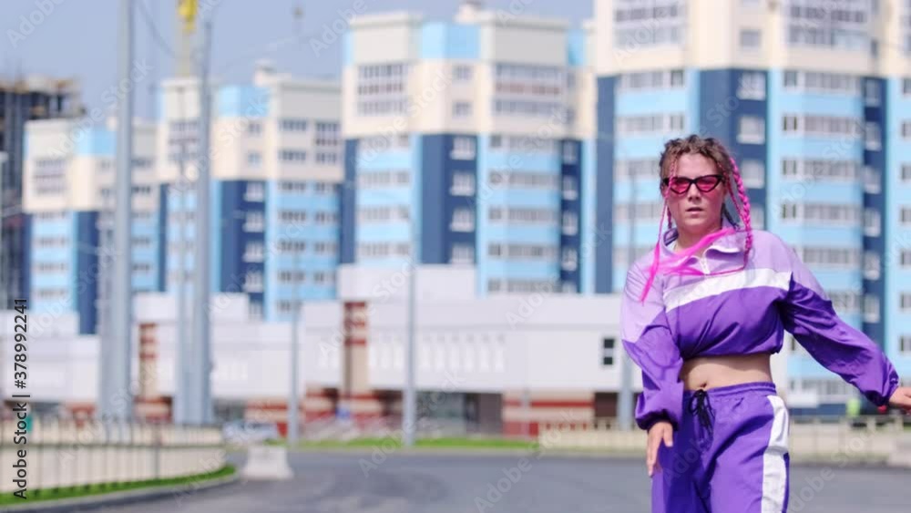 Funny girl with colored pigtails dancing hip hop outdoors on the background of high-rise buildings, jumping cheerfully and looking at the camera. Slow motion Funny girl with colored pigtails dancing hip hop outdoors on the background of high-rise buildings, jumping cheerfully and looking at the camera. Slow motion
