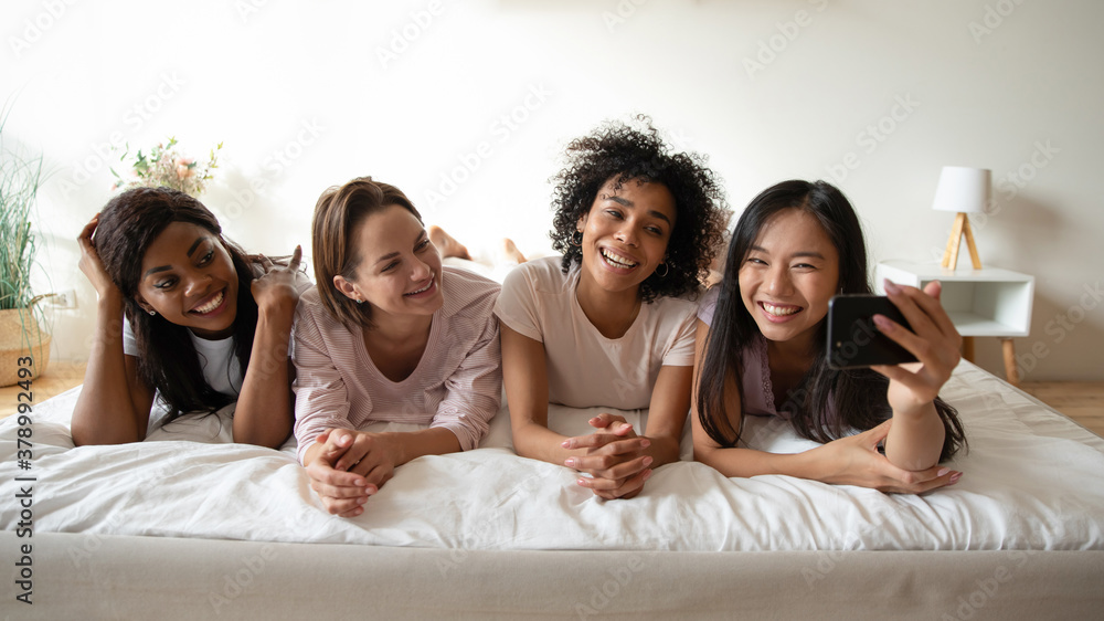 Overjoyed young beautiful smiling mixed race girls lying on comfortable bed, taking funny selfie photos or record video on smartphone, enjoying payamas sleepover party together, entertaining indoors. Overjoyed young beautiful smiling mixed race girls lying on comfortable bed, taking funny selfie photos or record video on smartphone, enjoying payamas sleepover party together, entertaining indoors.